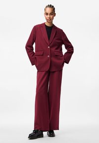 Burgundi ülikond kohandatud bleiseriga, millel on kaks taskut ja kaks nööpi, koos laiade pükstega. Mustad kingad täiendavad riietust.