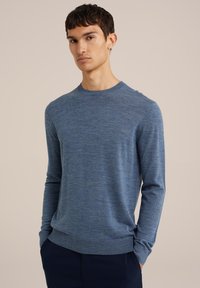 Kék kötött pulóver, lekerekített nyakkal, hosszú ujjal, finom textúrával, bordás mandzsettákkal és passzéval, sötétkék nadrág felett viselve.