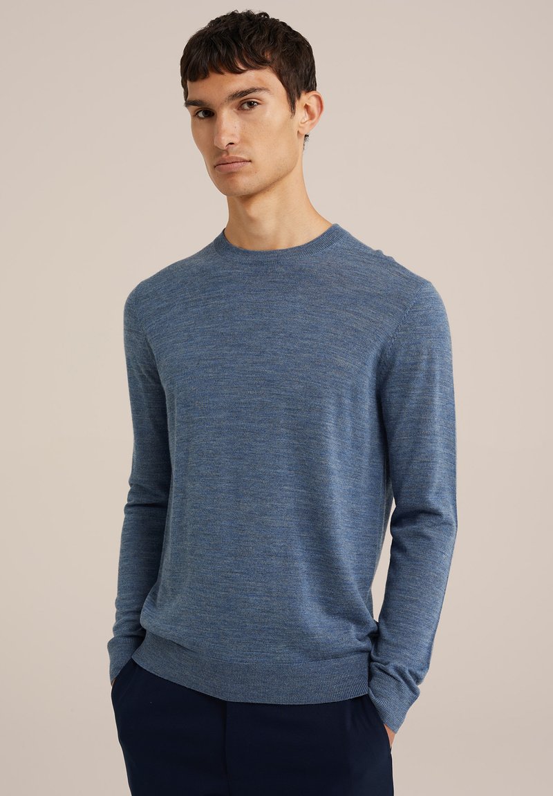 Kék kötött pulóver, lekerekített nyakkal, hosszú ujjal, finom textúrával, bordás mandzsettákkal és passzéval, sötétkék nadrág felett viselve.