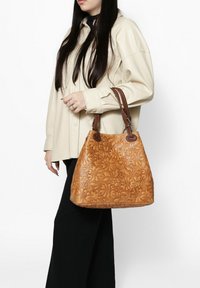 Tan reliëf handtas met bloemenpatronen, voorzien van bruine leren handvatten en een slouchy vorm, vastgehouden door een persoon in een crème shirt.