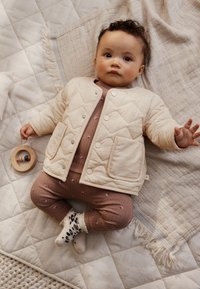 Veste matelassée beige avec boutons-pression, associée à un body rose côtelé avec de petites étoiles, et des chaussettes à motifs, sur une couverture texturée.