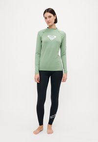 Grønn langermet rashguard med en hvit logo på brystet og ærmet, kombinert med svarte leggings med en hvit logo på ankelen.