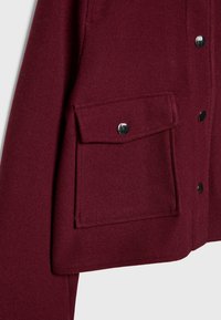 Gros plan d'une poche de manteau bordeaux avec un rabat sécurisé par un bouton-pression argenté sur un fond en tissu en laine.