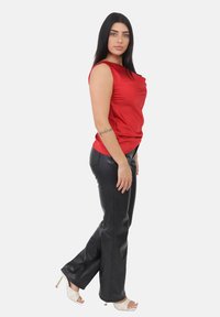 Vrouw met lang, donker haar, gekleed in een rode mouwloze top, zwarte leren broek, witte geweven hoge hakken en een tattoo op haar onderarm, staande in profiel.