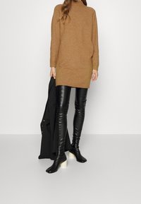 Svetr s vysokým výstřihem a dlouhými rukávy v barvě velbloudí, doplněný o přiléhavé černé kožené kalhoty a boty na vysokém podpatku s bledou podrážkou.