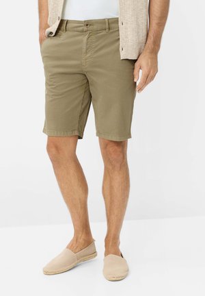 Mann trägt khakifarbene Shorts, beige Slipper, weißes Hemd und beige Strickjacke, die Hände in den Taschen, stehend auf weißem Boden.