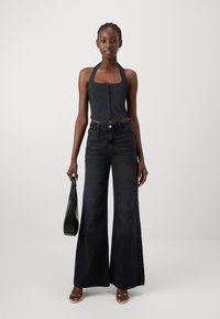 Črn halter top z detajli gumbov, v kombinaciji z visokim pasom in širokimi črnimi kavbojkami. Model drži črno torbico in nosi odprte čevlje.
