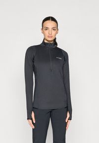 Sötét hajú nő lófarokban, fekete, hosszú ujj�ú, félzipzáras sportfelsőt és fekete nadrágot visel egy sima háttér előtt.