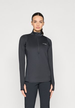 Femme aux cheveux foncés en queue de cheval, vêtue d'un haut de sport noir à manches longues et à zip demi-long, ainsi que d'un pantalon noir, sur fond uni.