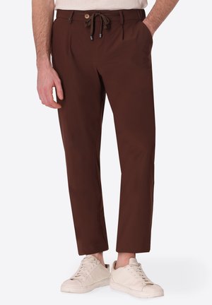 Uomo che indossa pantaloni marroni dritti con bottone e coulisse, abbinati a sneaker color crema e una mano in tasca.