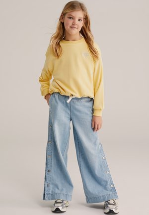 Žuta majica dugih rukava s okruglim izrezom vrata, uparen s širokim trapericama svijetloplave boje koje imaju detalje s gumbima sa strane. Ležerne atletske tenisice.