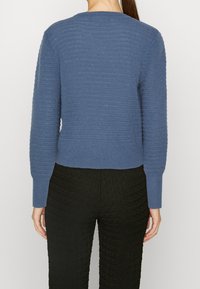 Blåtexturerad tröja med långa ärmar och ribbad nederkant, i kombination med svarta texturerade byxor. Bakifrån framhäver bilden den vävda mönstret på tröjan.