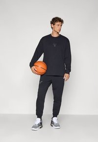 Musta pitkähihaispaita, jossa on hillitty logo, yhdistettynä sävyyn sopiviin collegehousuihin. Malli pitää kädessään koripalloa ja on pukeutunut harmaisiin tennareihin.