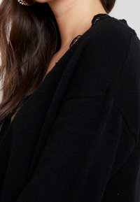 Schwarze Strickjacke mit tiefem V-Ausschnitt und zarter Spitzenverzierung an den Schultern. Weicher Griff und lockere Passform.