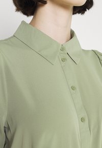 Kobieta ubrana w jasnozieloną koszulę z kołnierzykiem i krótkimi rękawami, pokazana od szyi do połowy torsu na białym tle.