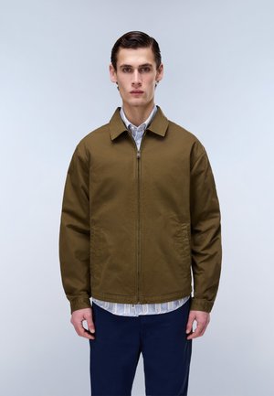 Ung mand stående med fronten mod kameraet iført en olivengrøn lynlåstrøje over en stribet skjorte og marineblå bukser mod en ensfarvet baggrund.