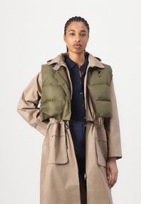 Gilet en duvet vert olive superposé à un trench-coat beige avec taille à cordon, doté d'un col et de deux poches avant.
