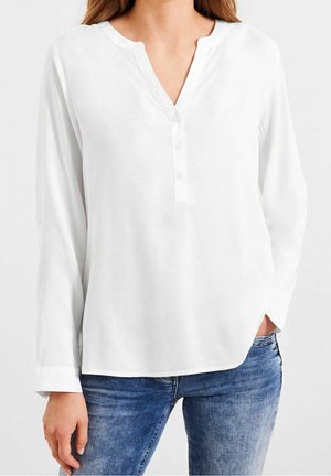 Vrouw draagt een wit blouse met lange mouwen en een V-hals met knopen, gecombineerd met een blauwe spijkerbroek, hand in de zak.