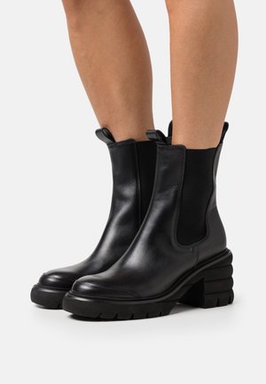 Black leather chunky-heeled ankle boots with elastic side panels worn on bare legs, shown against a white background.