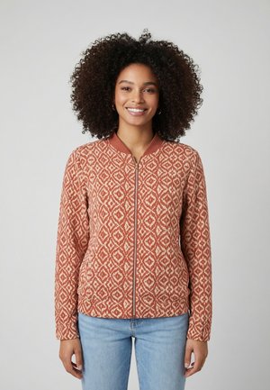 Femme souriante aux cheveux bouclés portant une veste zippée à motif rouille orange et crème, associée à un jean bleu clair, sur un fond uni.