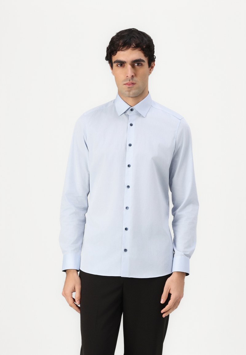 Chemise à manches longues bleu clair avec un col à boutons, dotée de boutons sombres et d'une texture de tissu lisse. Portée avec un pantalon noir.