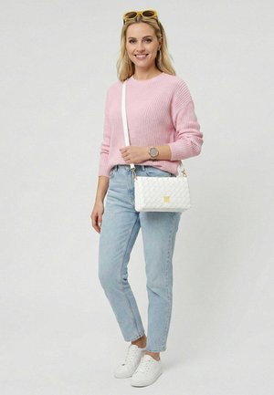 Femme portant un pull rose, un jean bleu clair, des baskets blanches et des lunettes de soleil jaunes posées sur la tête, portant un sac bandoulière blanc tissé.