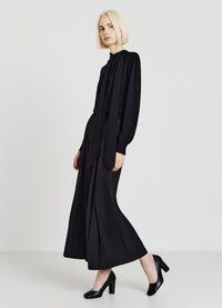 Schwarze Langarmbluse mit gerafften Schultern, kombiniert mit weitem Hosenbein. Modell trägt schwarze High-Heel-Schuhe. Glatte Stoffe, minimalistische Details.