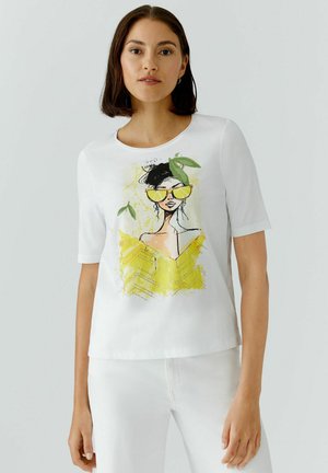 Frau trägt weißes Kurzarmshirt mit illustrierter Grafik einer Frau mit gelber Sonnenbrille und Kleid mit grünen Blättern auf hellem Hintergrund.