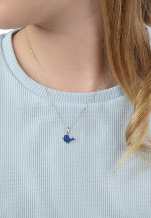 Collar de cadena de plata con un pequeño colgante de ballena azul, llevado sobre una camiseta azul claro de canalé, parcialmente cubierto por cabello rubio.