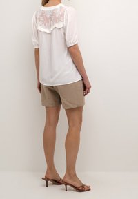 Blusa blanca con yugo trasero bordado translúcido, mangas abullonadas y dobladillo con volantes, combinada con pantalones cortos beige y sandalias de tacón marrones.