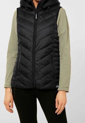 Person iført en sort quiltet ærmeløs vest med hætte over en langærmet beige og sort stribet skjorte og sorte bukser.