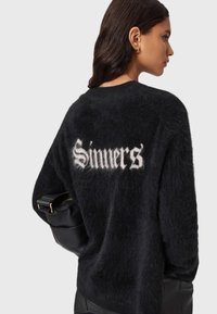 Musta pörröinen neule rentolla leikkauksella, jonka selässä on suuret valkoiset sanat "Sinners'". Materiaali näyttää pehmeältä ja villaiselta.