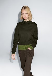 Dunkelgrüner, gekürzter Pullover mit einem Schalkragen, kombiniert mit einem hellgrünen Langarmshirt darunter und braunen Hosen. Glatte Textur.
