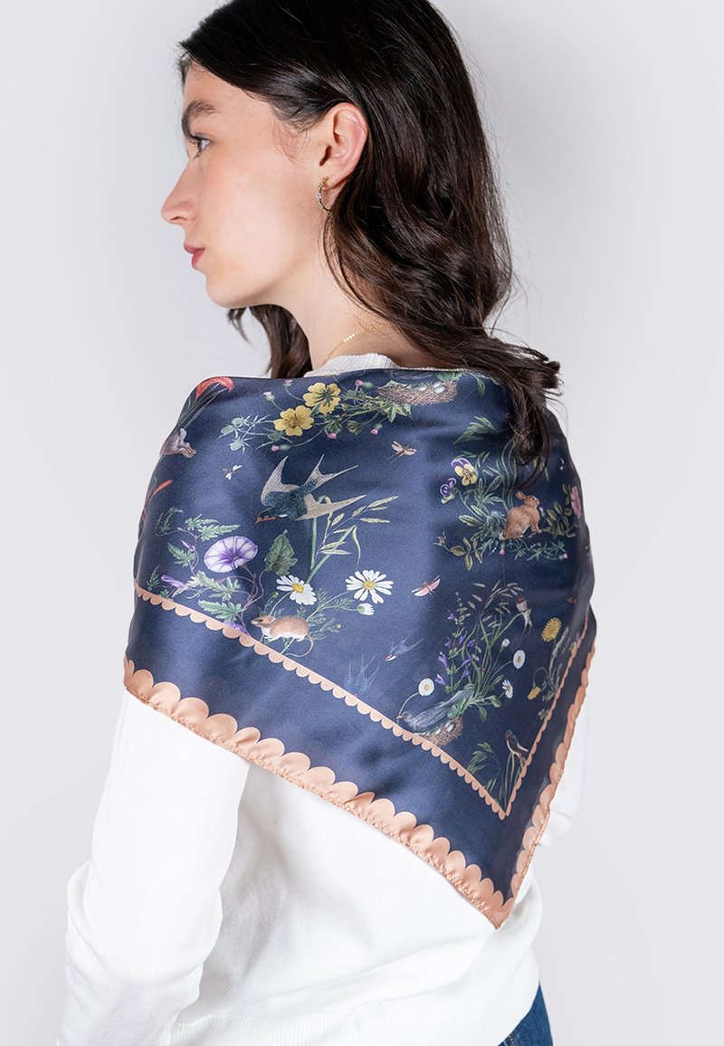 Écharpe en soie avec un motif floral et animal en bleu marine, ornée de bords en cuivre festonnés. Drapée sur l'épaule contre un haut blanc.