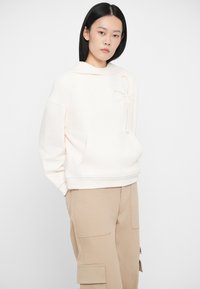 Ženska s ravnimi črnimi lasmi, oblečena v off-white kapuco s žepom in bež cargo hlače, stoji proti enostavni svetli ozadju.