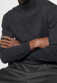 Un pull à col roulé gris foncé en tissu tricoté texturé, avec des manches longues et des poignets côtelés, associé à un pantalon sombre.