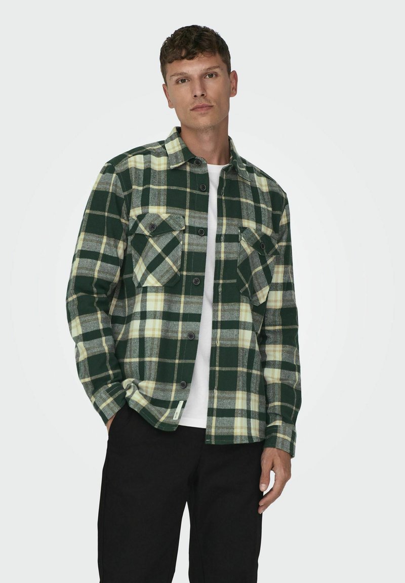 Camisa de franela a cuadros en verde y crema con dos bolsillos en el pecho, cierre de botones en el frente y mangas largas, llevada sobre una camisa blanca.