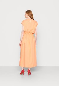 Vestido maxi laranja sem mangas com cintura franzida, detalhe em buraco na parte de trás, fendas laterais e usado com sapatos de salto alto vermelhos.