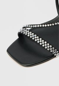 Czarne sandały ze kwadratowym noskiem, z krzyżującymi się paskami ozdobionymi przezroczystymi cyrkoniami dla dodatkowego efektu. Gładka, skórzopodobna tekstura.