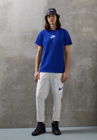 Blå bomullströja med vitt Nike-logotyp, vita cargobyxor med blå Nike-swoosh, svarta hög-top sneakers och en svart bucket hat.