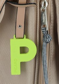 Grünes Kunststoffschriftzug "P"-Etikett, das an einer beigefarbenen strukturierten Tasche befestigt ist, mit einem silbernen Reißverschluss und einem schwarz-weißen karierten Kordeldetail.