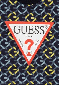 Pamučna majica s tamnoplavom pozadinom i geometrijskim uzorcima u plavoj, žutoj i sivoj boji. Crveni trokutasti logotip s istaknutim natpisom "GUESS".