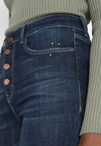 Mörkblå denimjeans med hög midja, femknappsflygt, subtilt slitage och detaljer i guldton. Bomullstyg med lätt stretch.