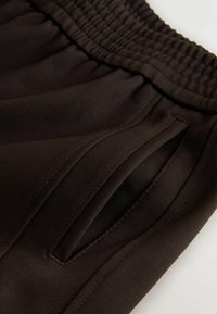 Close-up of dark brown pants waistband with elastic band and side slit pocket showing detailed stitching and fabric texture.