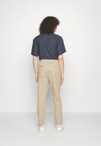 Námořnicky modrá košile s krátkým rukávem, béžové kalhoty na míru, hladká textura, úzký střih, s klasickým límcem a zadními kapsami, doplněné bílými botami.