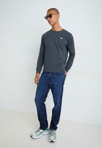 Camiseta gris de manga larga con un pequeño logo, combinada con jeans de mezclilla azul y zapatos deportivos coloridos. Modelo con gafas de sol.