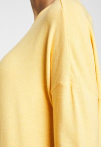 Gelbes strukturiertes Sweatshirt mit lockerer Passform, abfallenden Schulternähten und einem glatten Strickmuster. Zeigt einen weichen, leichten Stoff.