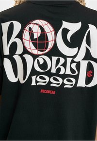 Juodas marškinėlis su ryškiu baltu ir raudonu tekstu "ROCA WORLD 1999" ir raudona žemės rutulio grafika, tekstūruotas medvilnės audinys, laisvas kirpimas.