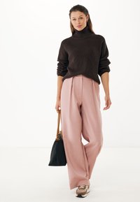 Jersey de cuello alto de canalé marrón, pantalones plisados rosa claro, zapatillas marrón y beige, y un bolso negro con correas marrones. Fondo blanco.