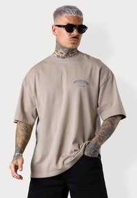 T-shirt oversize beige clair avec des manches courtes, un col rond et un accent imprimé sur la poitrine, associé à un pantalon noir et des lunettes de soleil.
